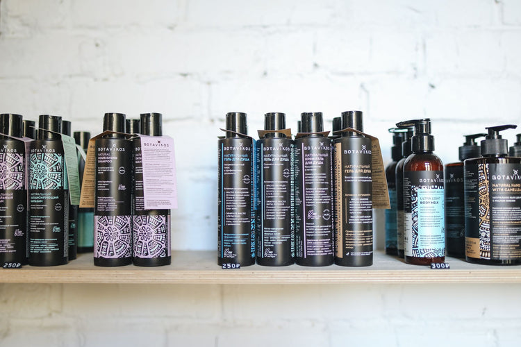 A variety of natural cosmetic bottles lined up on a wooden shelf against a white brick wall, suggesting eco-friendly bea